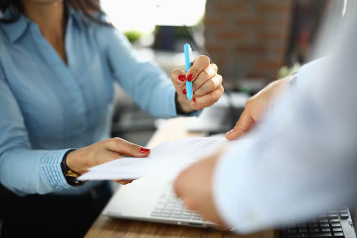 A businesswoman signs a document that’s handed to her while working with a laptop.