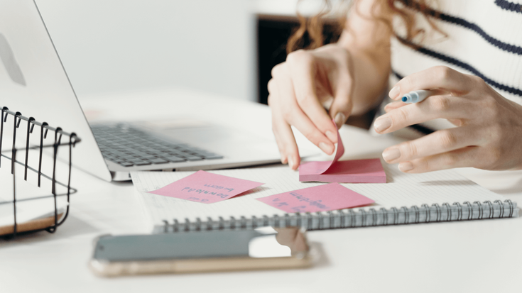 A woman writes sticky note reminders beside a laptop and smartphone.